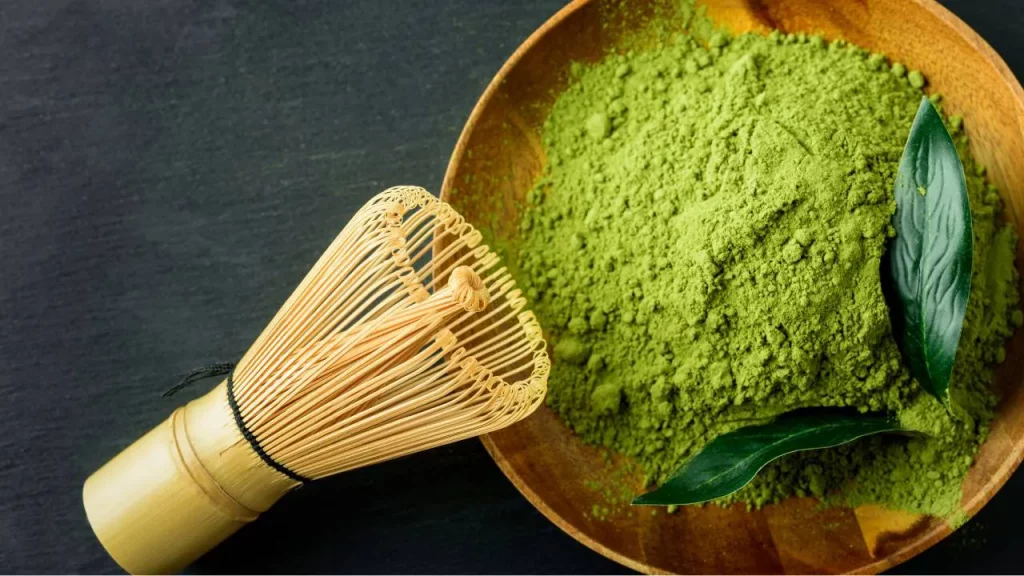 Bol en bois contenant de la poudre de thé matcha vert avec un fouet en bambou chasen et des feuilles de thé fraîches